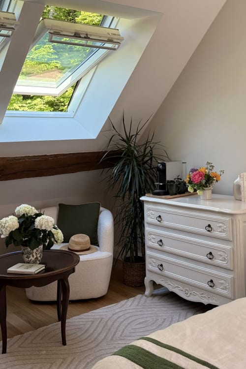 Bright attic bedroom at La Maison 1680 with skylight, cream armchair, white dresser, and garden views in Couches, France