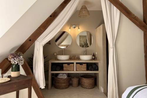 Attic bathroom at La Maison 1680 with dual vessel sinks, round mirrors, exposed wooden beams, and woven storage baskets