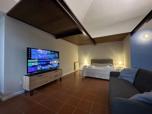 Modern loft bedroom at Cinelli Apartment in Vercelli with 75-inch TV, queen bed, dark wood ceiling beams, and terracotta tile