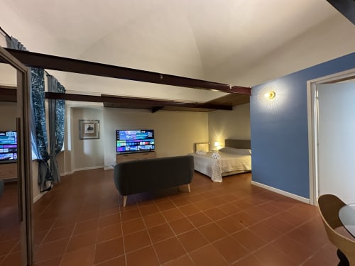 Spacious loft apartment in Vercelli with terracotta flooring, exposed beams, blue accent wall, TV, and bedroom visible in