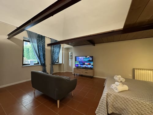 Modern loft living room with black armchair, 75" TV, wooden beams, and bed with towels in Vercelli historic center