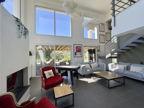 Salon de villa moderne avec fauteuil en velours rouge, canapé gris, vue sur la piscine par les baies vitrées et escalier mezzanine