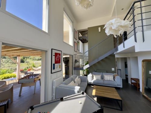 Salon de villa moderne avec hauts plafonds, luminaires suspendus blancs, canapés gris et vue sur la terrasse jardin à SoleMare,
