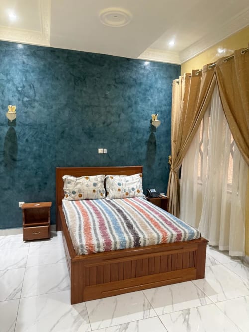 Chambre double de luxe au Kylian Appart Hotel, Brazzaville avec lit en bois, mur d'accent bleu et rideaux donnant sur le jardin