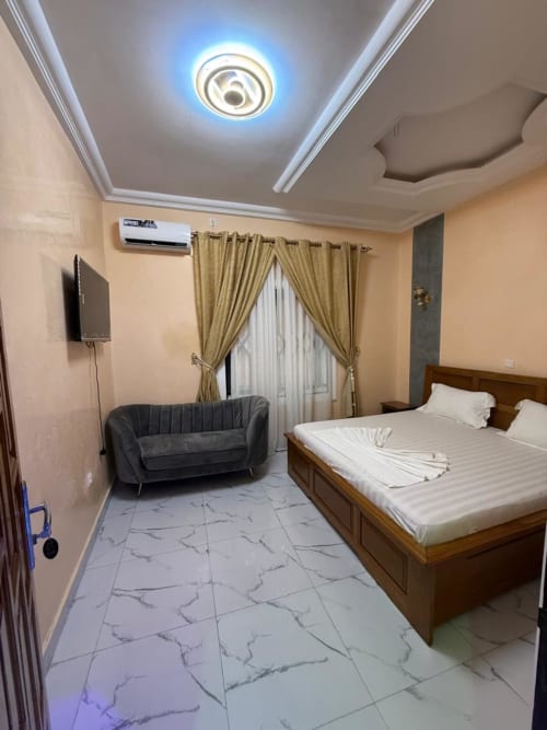 Chambre double luxe au Kylian Appart Hotel de Brazzaville avec lit en bois, canapé gris, climatisation, TV écran plat et carrelage en marbre