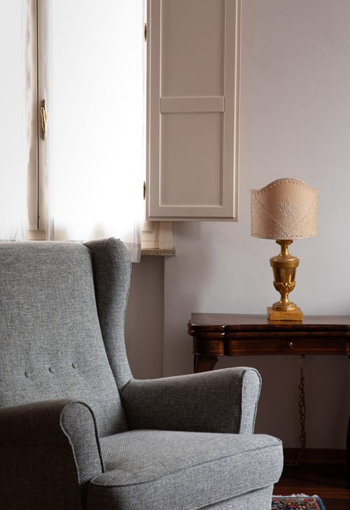 Gray wingback chair by window in Cristallo Apartment, Ravenna hotel room with lamp and wooden desk