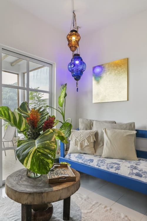 Bright cottage interior with blue hanging lamp, tropical flowers, wooden round table, and blue bench seating with sea views