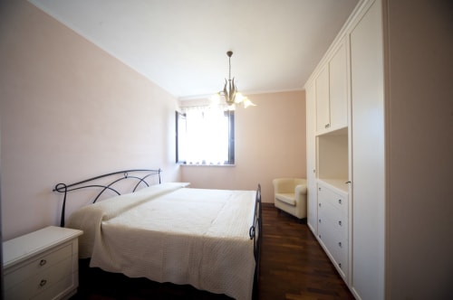 Chambre spacieuse et lumineuse avec lit en fer forgé, couvre-lit crème, fauteuil crème, armoire blanche et parquet