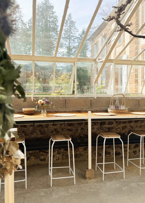 Salle à manger de la serre Maison Mansel avec comptoir en bois, tabourets de bar et vue sur le jardin à Mesnil-en-Ouche
