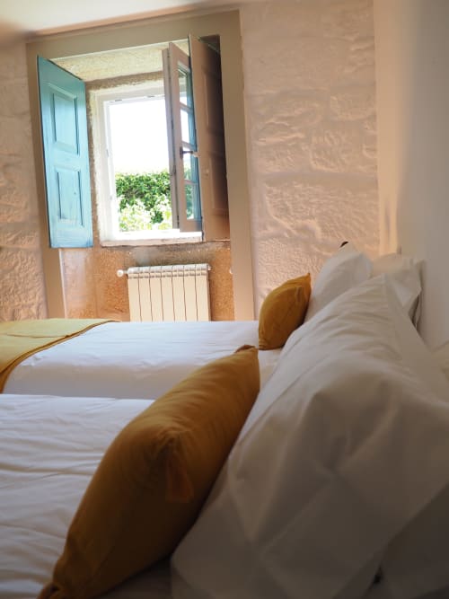 Quarto acolhedor na Quinta da Coutada com janela de venezianas azuis, roupa de cama branca e almofadas douradas em Arcos de Valdevez