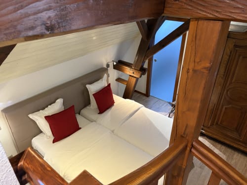 Chambre spacieuse à l'Hôtel de l'Ange avec lits jumeaux, oreillers rouges, poutres en bois et vue sur le jardin