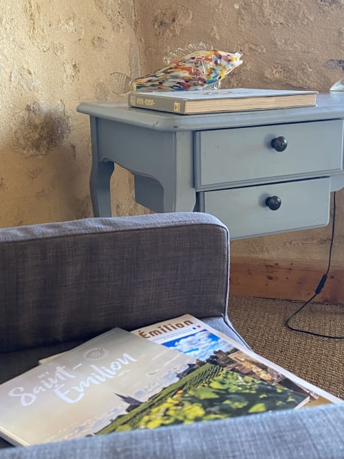 Cozy bedroom corner at Le Prieuré de Saint-Pierre with gray nightstand, magazines, and rustic stone wall with garden view.