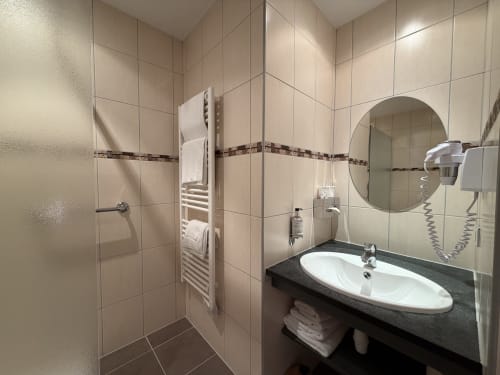 Salle de bains moderne de l'Hôtel de l'Ange : lavabo blanc, miroir rond, radiateur sèche-serviettes et douche à l'italienne.