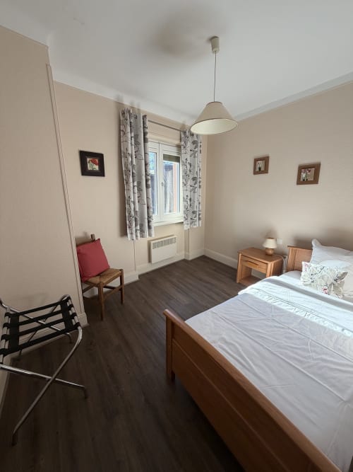 Chambre spacieuse à l'Hôtel de l'Ange avec lit en bois, bureau, chaise, suspension et fenêtre sur jardin avec rideaux
