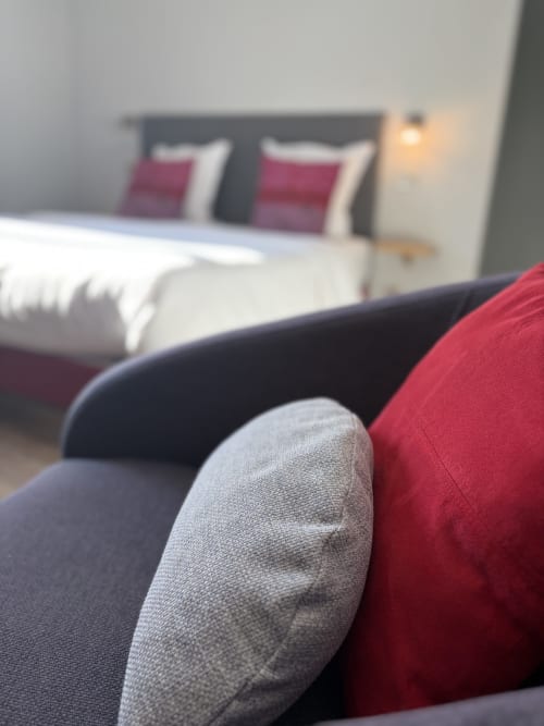 Chambre moderne de l'Hôtel de l'Ange avec lit double, accents bordeaux et assise grise à Niedermorschwihr