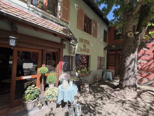Entrée de l'Hôtel de l'Ange à Niedermorschwihr avec plantes fleuries, table bleue et charmante cour par une belle journée ensoleillée