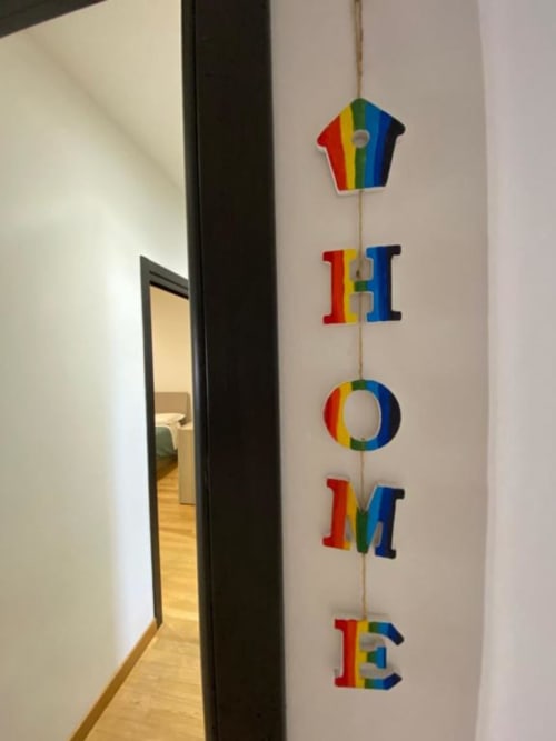 Insegna colorata 'HOME' con lettere arcobaleno appesa al muro bianco vicino all'ingresso degli MiSaNi Apartments