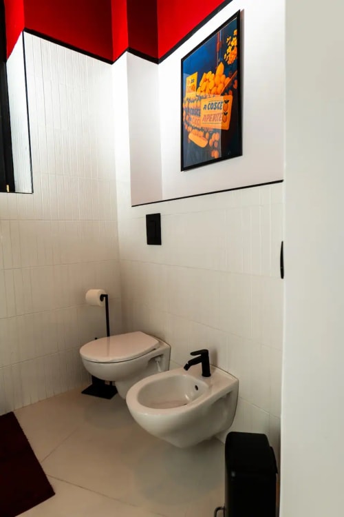 Bagno moderno dell'appartamento LH51 a Napoli con piastrelle bianche, toilette, bidet, rubinetteria nera e poster vintage su parete rossa