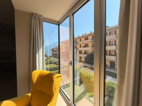 Bright yellow chair by floor-to-ceiling windows overlooking Italian architecture and courtyard at DV Garda Suite, Campione
