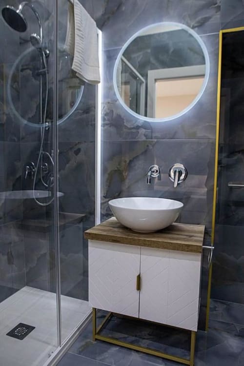 Bagno moderno a Dreams & Colors Napoli con specchio circolare illuminato, lavabo bianco in ceramica, rivestimenti in marmo e pareti in vetro