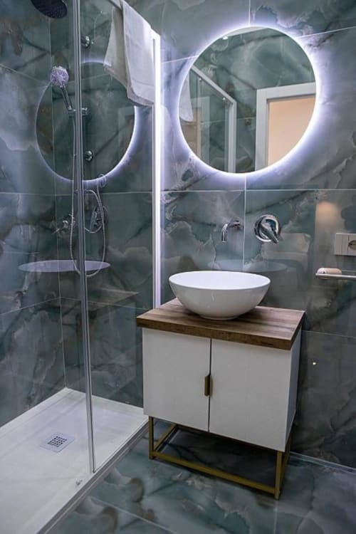 Bagno moderno a Dreams & Colors Napoli con specchi LED doppi, lavabo bianco a coppa, pareti in marmo, doccia in vetro e legno