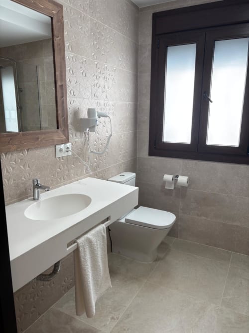 Modern bathroom in Hotel Mulhacén Trevélez suite with white sink, toilet, textured beige tile walls, and dark-framed windows