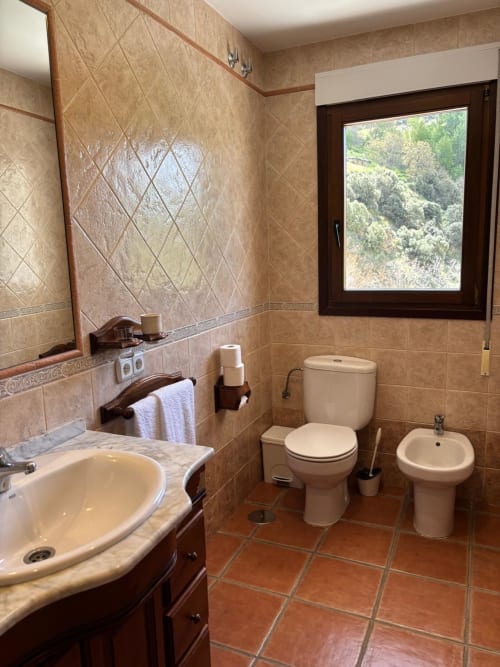 Bathroom with beige tiled walls, sink, toilet, bidet, and window overlooking forested hillside at Hotel Mulhacén Trevélez,