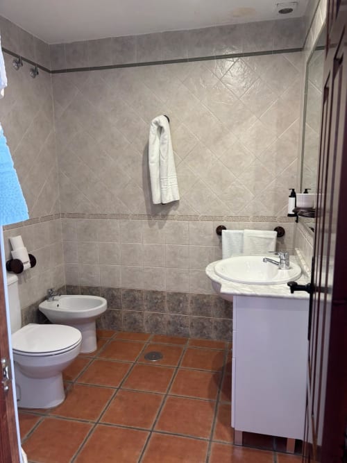 Baño moderno del Hotel Mulhacén Trevélez con lavabo blanco, bidé, inodoro, paredes alicatadas y suelo de barro