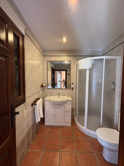 Baño moderno del Hotel Mulhacén Trevélez con ducha curva, lavamanos blanco, inodoro y suelo de baldosas de terracota