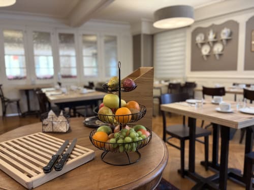 Présentoir à fruits à trois étages sur table de restaurant à l'Hôtel de l'Ange avec couverts et fenêtres.
