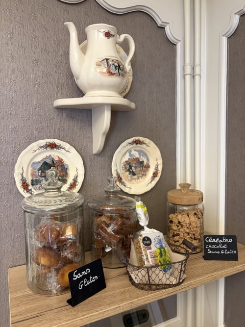 Étagère décorée à l'Hôtel de l'Ange présentant une théière en porcelaine vintage, des assiettes ornées et des en-cas sans gluten en pots de verre.