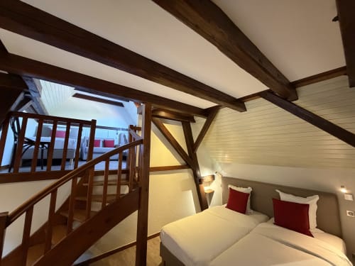 Chambre cosy avec poutres en bois, draps blancs, oreillers rouges et escalier mezzanine à l'Hôtel de l'Ange