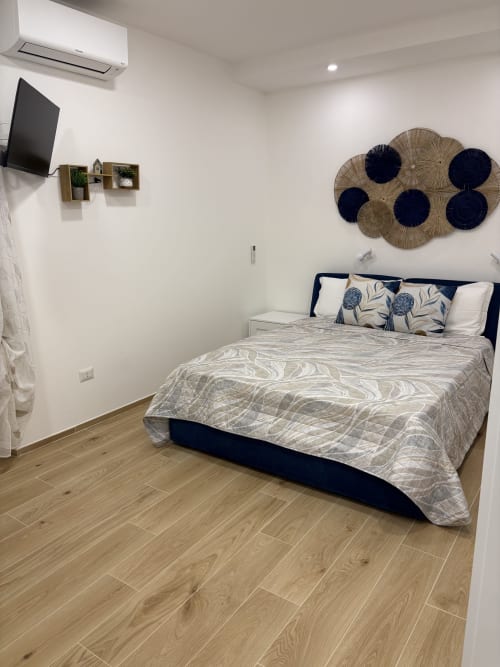 Modern bedroom at Catalogna10 Rooms in Alghero with queen bed, navy headboard, woven wall art, TV, AC unit, and wooden