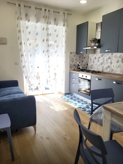 Modern kitchen-living area in Alghero apartment with gray cabinets, wooden floors, and garden view through sheer curtains