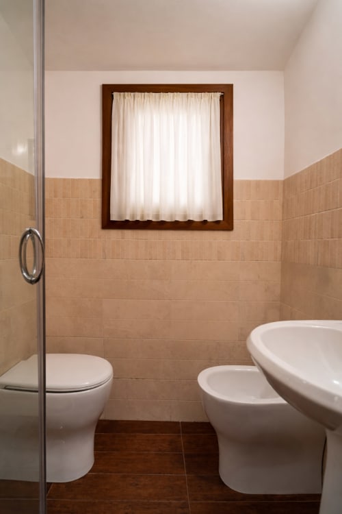 Bagno moderno con piastrelle beige, sanitari bianchi, bidet e finestra con tende presso il cottage Stone House a Novi Velia