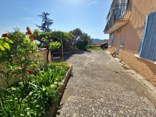 L'allée du Smooth Luberon à Villars avec ses roses rouges en fleur, bâtisse en pierre aux volets bleus et vue sur les montagnes