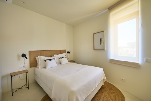 Bright bedroom at Casa Redonda Aroeira in Portugal with queen bed, woven rug, wooden nightstand, and pool view window