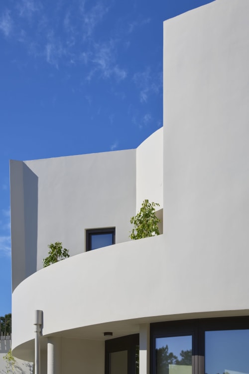 Modern white curved villa at Casa Redonda Aroeira with blue window and green plants against clear sky
