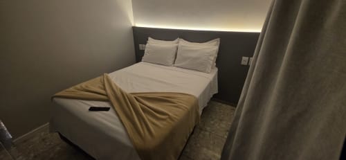 Suite moderna no Downtown Rooftop 375 em Macaé com cama queen, roupa de cama branca, cabeceira escura, iluminação ambiente e cortinas translúcidas