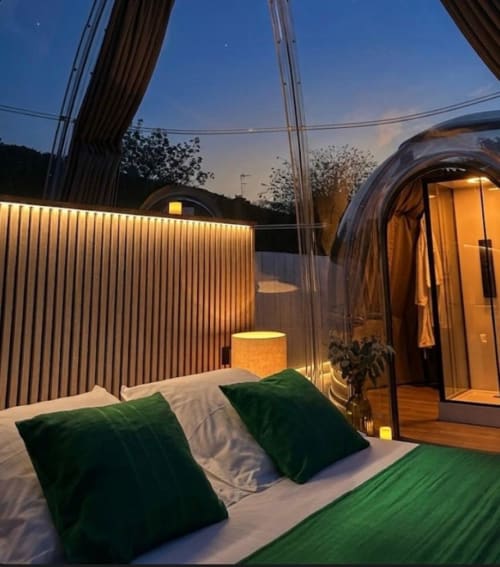 Chambre Bubble Jade à la tombée du jour avec lit illuminé, coussins verts, structure de pod en verre courbe et patio privé à Bazouges
