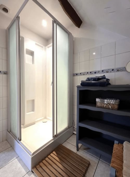 Modern bathroom with glass shower enclosure, white tiles, and dark shelving unit at La Sauge room, Ferme de la Forterie