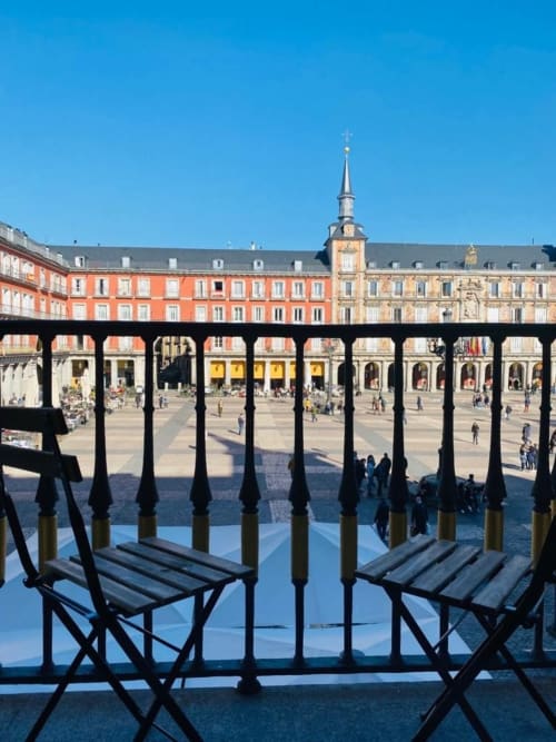 Black chairs on covered balcony overlooking Plaza Mayor Madrid with red buildings and church tower under blue sky