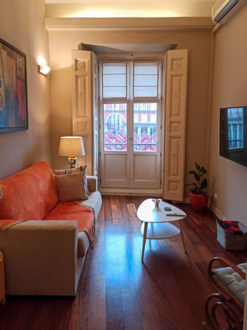 Modern studio apartment at Plaza Mayor with orange sofa, wooden floors, balcony doors overlooking city, and contemporary
