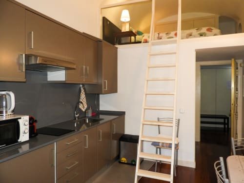 Studio apartment kitchen with brown cabinetry, black countertops, and wooden loft ladder leading to sleeping area in Madrid.