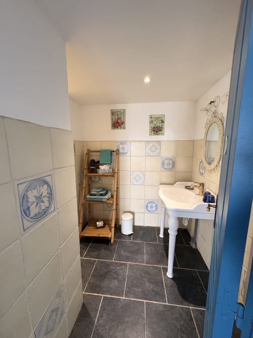 Bathroom at Ferme de la Forterie B&B in Verlinghem with pedestal sink, decorative blue tiles, wooden shelf storage