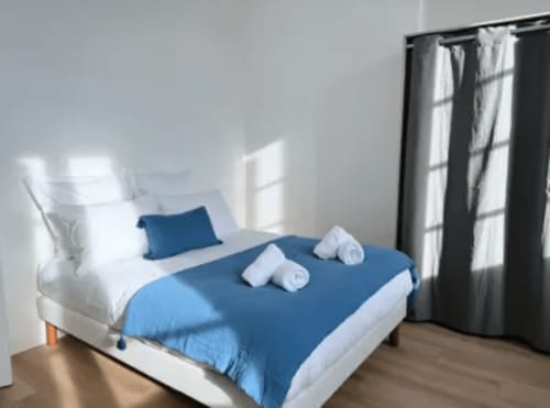 Chambre moderne de l'hôtel ARLOÉ à La Rochelle avec literie bleue, oreillers blancs, rideaux sombres et parquet