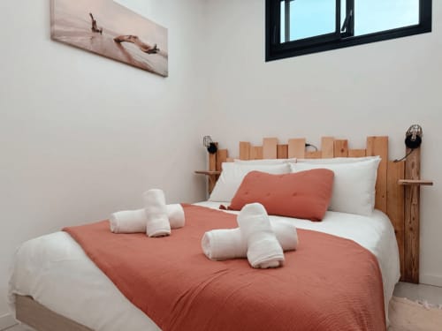 Chambre douillette de l'appartement ARLOÉ à La Rochelle avec tête de lit en bois rustique, couvre-lit corail, oreillers blancs et serviettes roulées