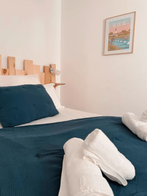 Chambre cosy d'ARLOÉ La Rochelle avec literie bleu-vert, tête de lit en bois, serviettes blanches et art mural côtier