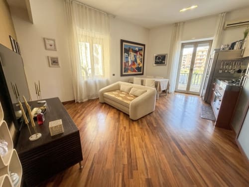 Luminosa e spaziosa zona living di ROOMITO APARTMENTS a Napoli con divano color crema, pavimenti in legno, cucina moderna e vista sulla città