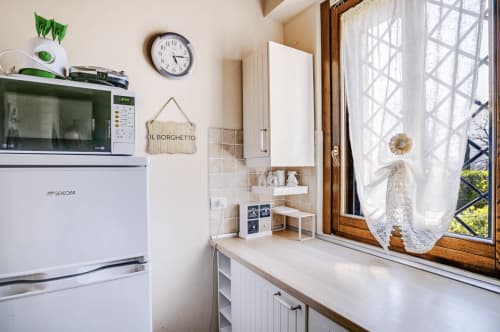 Compact kitchen at Casa Rossi Roma with white appliances, wall clock, green plants, and window overlooking Rome